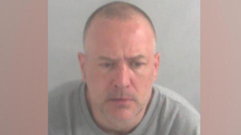 A police custody photograph of Jeremy Heath in front of a grey backdrop. He is wearing a grey round-necked jumper. He has close-shaved dark greying hair and stubble