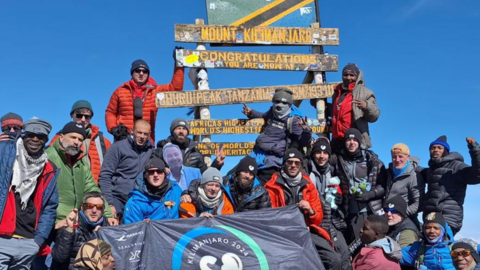 The group of 20 climbers from the Strongmen charity who climbed Kilimanjaro together. Many are wearing hats and sunglasses and warm coats, and they are posed around signs at the peak of the mountain.