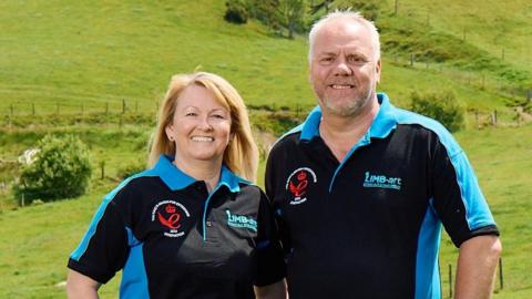 Mark Williams stands in a field alongside his wife, Rachael. Mark has close-cropped grey hair and a short beard and he's smiling into the camera. Rachael has blonde hair and is smiling too. Both of them are wearing black and blue T-shirts with their company's logo.