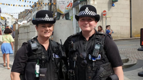 Two female police officers in uniform are standing in a street and smiling at the camera. They both have their hair tied back and are wearing police hats. They have handheld radios and cameras on their heavy duty waistcoats along with sunglasses and hand sanitizer gel.