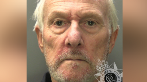 A police custody image of Peter Rowell. He is in his 60s with messy grey hair and short grey stubble. The collar of a dark shirt is visible. Gwent Police's Logo sits in the bottom right-hand corner. 