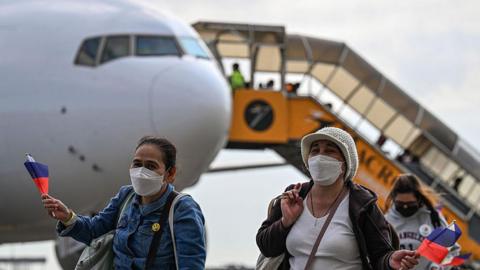 Three women waving the Philippine flag and wearing face masks, walking from an airplane in the background after being repatriated fro the Middle East due to the US and Israel's war on Iran