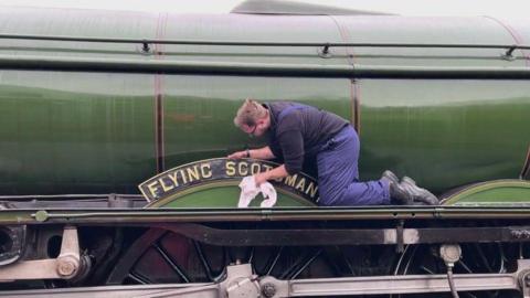 A man wearing blue work overalls polishes a sign reading 'Flying Scotsman' with a white drag. It is attached to the side of the large green train. 