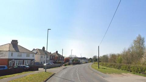 A street view of Moira Road taken from the middle of the road. On the left are three pairs of semi-detached houses, cars parked on the drives, and a pavement next to the road. On the right side is a pavement, short hedge and trees.