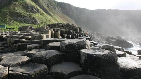 Giants Causeway - several dozen dark, circular rocks. Behind the rocks, there is a large cliff-face with plenty of grass. On the right, the sea is meeting the rocks. It is a sunny day.