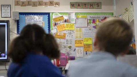 Classroom wall display with ‘Spelling’ and ‘Maths’ labels, covered in student work, notes, and diagrams, with two students visible out of focus in the foreground