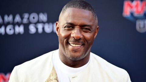 Luther Idris in a cream jacket and white T-shirt