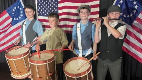 Four young boys stand in front of American flags. 3 of them have large drums and are holding drumsticks. The fourth has a flute to his lips. They are dressed in told fashioned clothes with waistcoats and flat caps. 