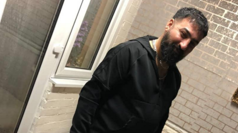 Mohammed Ali Nareman, a man with dark hair and a large beard, poses for a photograph wearing a black hoodie.