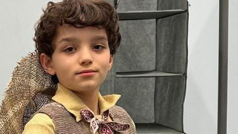 Tyne, wearing a Munchkin costume, looking at the camera. He has short dark hair, has a hat on his back, a yellow shirt, tie round his neck, and brown jacket. A rail is to his left. 