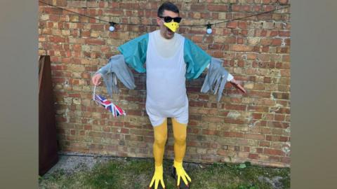 Sam from Rochester dressed as a seagull. Sam is wearing a white overshirt, yellow tights, yellow claw shoes and a beak. He is holding a Union Jack flag.