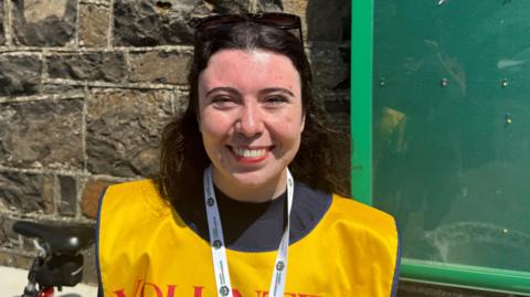 Keely is smiling. She has brown hair. She's wearing a yellow bib that says volunteer. She's outside.