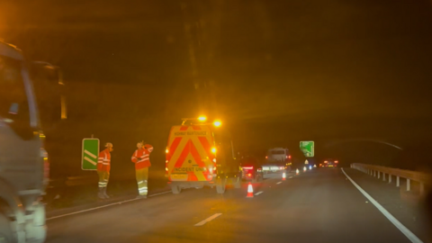 A highway maintenance vehicle is stopped on a dual carriageway. Traffic cones have been placed on the road and two men in high vis are standing at the side of the road talking to each other. 