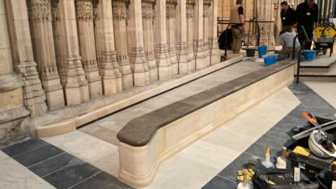 A long, carved stone ramp runs alongside an ornate wall with detailed vertical columns and decorative patterns. Several workers are installing or repairing the flooring, using buckets, tools, and equipment spread across the area. The setting appears to be a historic or grand indoor space, possibly undergoing restoration or maintenance.