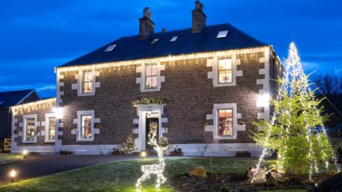 A large, two storey farmhouse, adorned with warm white Christmas lights. In the front garden there is a light-up reindeer and a large Christmas tree decorated with fairy lights. A wreath sits on the front door. 