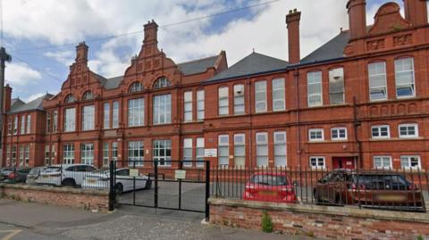 A red brick school building of three stories, with a car park in front and several cars parked there. There are railings around it which are closed.