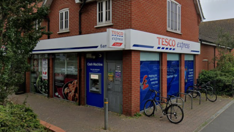 A general view of Tesco Express of Nacton Road in Ipswich. It shows the store front with the Tesco Express branding. A bicycle rack can be seen as well as a tree and bushes. A cash point is on the exterior of the building.