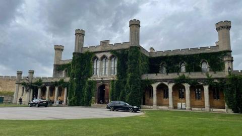 Exterior of Lincoln Crown Court a two-storey Victorian Gothic stone building with crenellated towers and vegetation growing around the door an upper windows. There is a grass lawn outside and two cars are parked outside on the paved forecourt.