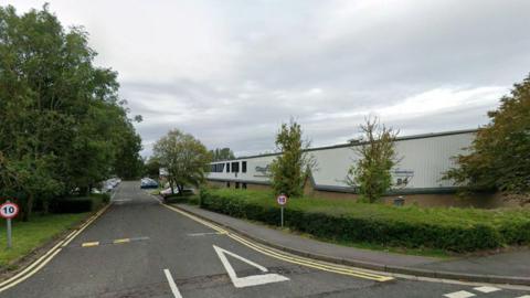 A factory building sits on the right hand side of the road lined with bushes and trees. 