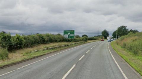 A large road, on approach to a roundabout, with a large green road sign on the left of the image.