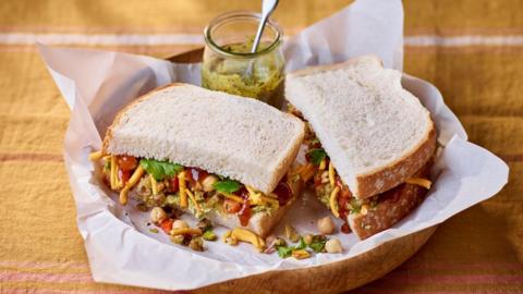 A filled sandwich cut in two sitting in baking paper on a plate. Alongside it is a small jar of chutney