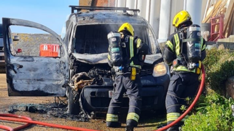 Two fire officers putting out a fire on an electric van, which is melted at the front and charred from the fumes.