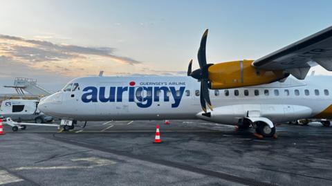 A close up side view of an Aurigny aircraft. The plane is white and yellow. Aurigny is written down the side. 
