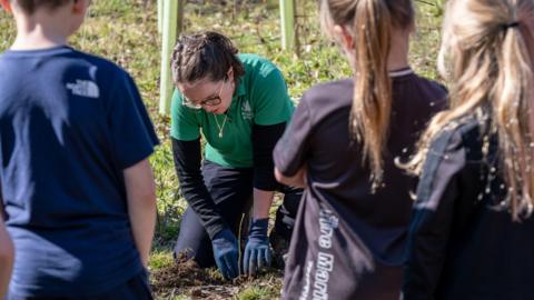 In the foreground are the backs of three children, a boy and two girls, in casual sportswear. They are standing watching a woman knelt down in the background, on grassy ground, tending to a sapling just planted into the ground. The woman is wearing a long sleeve black t-shirt, with a short sleeve green Forestry England branded polo top over the top of the t-shirt. She is wearing black trousers, blue gardening gloves, and she has brown hair tied up and is wearing glasses. It's sunny.
