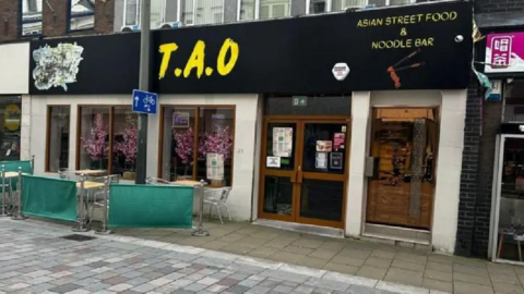 Front face of restaurant TAO. The signage is black with yellow print, and the window has pink flowers as decoration. There are tables and chairs outside the site, which are empty.