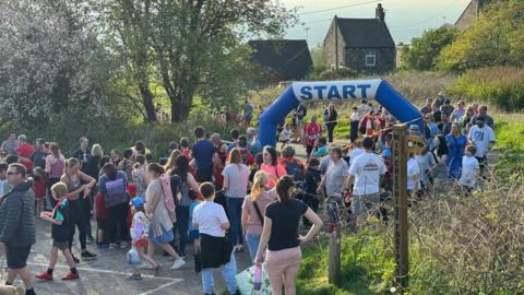 A large number of people in a rural village standing next to and walking through a start/finish line. A blue and white inflatable archway says "start" in the middle.  