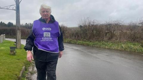 Derrick Downs wearing a mauve bib which says Oasis. He is walking along the side of a road towards the camera in wet conditions.