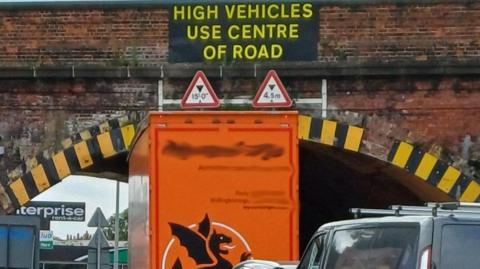 A orange lorry is trapped under a red-brick arched bridge, which is highlighted by yellow and black striped reflective signs. Triangular signs denote the height and a warning reads: "High vehicles use centre of road".