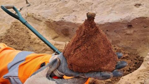 A close up of two gloved hands holding a sand-covered shield boss, with a wide base narrowing to point on the top. Behind it is an excavated grave in the sandy soil.