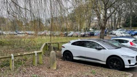 A car park full of cars with a grassy area and trees in the middle. 