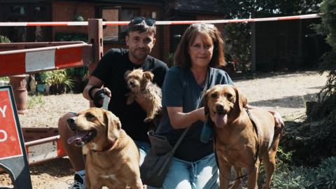 Aaron Fulcher crouching on the ground in front of his mum's former home. Next to him is Siobhan Fulcher. The pair of them have three dogs, two are on leads and one is being held in Aaron's arm. They are both crouched on the ground and are looking directly at the camera.
