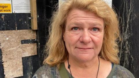 A woman with long blonde hair and wearing a green top is stood in front of a black door that has paint peeling away from it. 