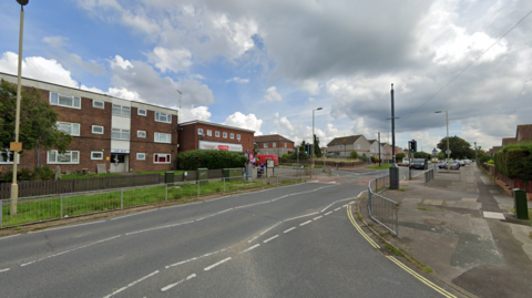 A generic Google image of Middle Park Way -  a wide junction with footpaths  with a three-storey block of flats opposite, a convenience store and houses further down the street