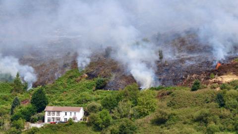 White smoke coming out of fires on a green valley. There is a white house with a terracotta roof in the foreground of the photo
