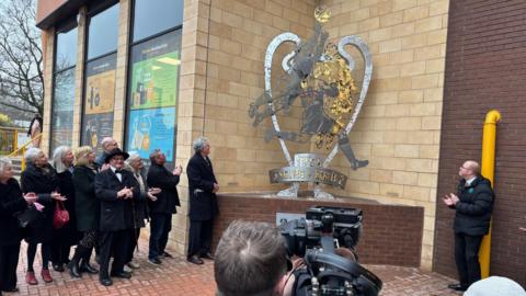 A group of people gather round a statue, they appear to be clapping. The statue is gold and silver in appearance and is shaped like two handles on a trophy with an inscription below and imagery of footballers in the centre.