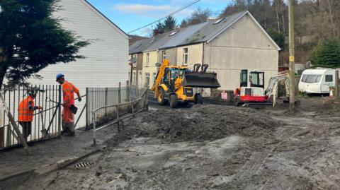 A street in Cwmtillery covered in black slugged piled to make it impassable. Two men in high vis orange jumpsuits and hard hard carry shovels as they stand on the pavement. A yellow digger with a large scoop and a smaller white and red digger with a small scoop are working to move the debris 