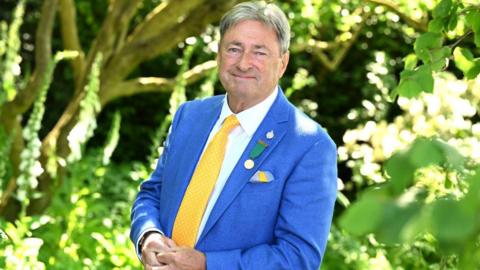 Alan Titchmarsh standing in a garden wearing a light blue suit, white shirt and yellow tie, a few medals are pinned to his lapel.