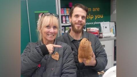 Two Morrisons staff - a man and a woman - stand with a cuddly toy capybara behind a kiosk. The lady is pointing at the toy.