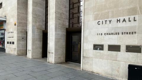 The exterior of the entrance to City Hall in Leicester, the City Council's headquarters. It has a light stone wall and three arches with black doors.