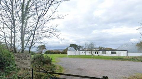 A white sign to the left with writing on it. The sign is amongst bushes and behind it is on the field. There is a white building on the right with windows. The sky is blue with white clouds.