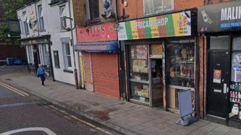 A pavement runs alongside Newport Street in Bolton, on which there are a number of shops, including African good shops Maje Worldwide. Food can be seen in the window as well as wigs. A woman is walking along the street with a shopping bag. 