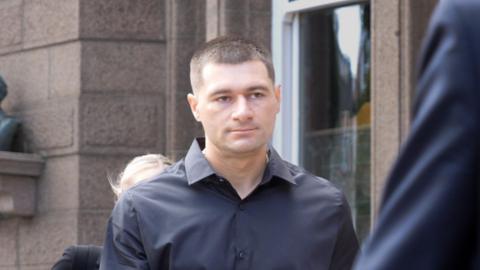 Artur Sevash-Zade wearing a black shirt walks into court. He has short black hair and is clean shaven.