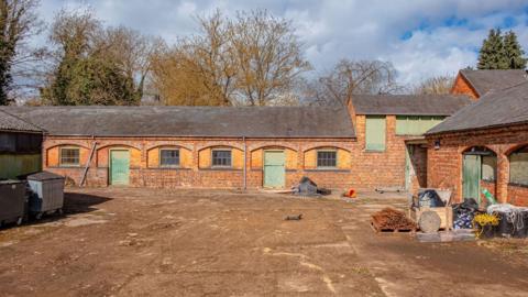 A dusty yard with a red-brick stable building with green doors in the backdrop.