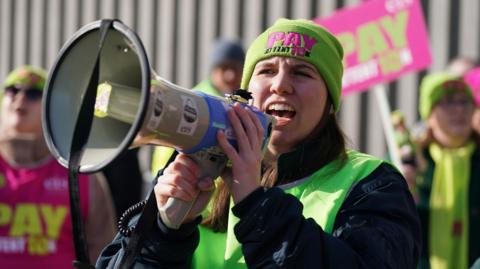 A female union member speaking animatedly into a megaphone. She has long brown hair and wears a lime green hat with the slogan "pay attention". 