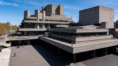 The National Theatre on London's South Bank is a a grey concrete building with stepped terraces, sat under a blue sky