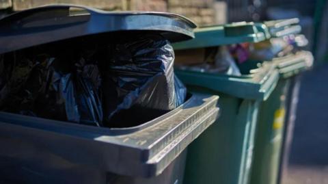 A black wheelie bin is in the foreground with the lid slightly open, exposing a black bin bag. Next to it, in the background, are two green recycling bins, also with the lids slightly open and exposing rubbish.